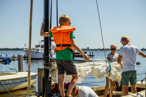 Children and adults wearing life jackets prepare a sailboat on a sunny day at a lake in Friesland, Netherlands.