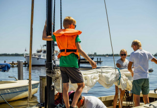 Børn og voksne iført redningsveste gør en sejlbåd klar på en solrig dag ved søen i Friesland, Holland.