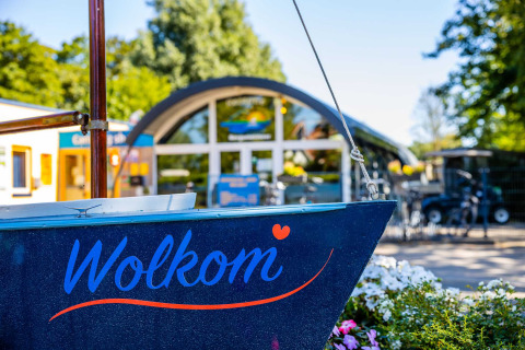 Entrada al Vakantiepark Bergumermeer, Frisia, Países Bajos, con un bote que dice 'Welkom' y flores delante.