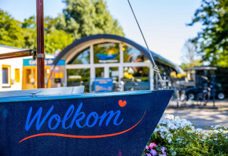 Indgang til Vakantiepark Bergumermeer med en båd, hvor der står 'Welkom' og farverige blomster i forgrunden.