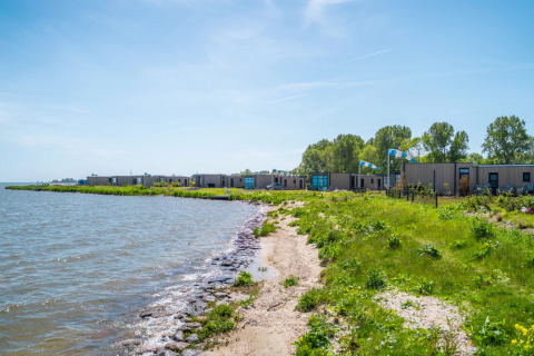 Parque vacacional junto al lago con alojamientos glamping modernos, vegetación verde y cielo azul claro.