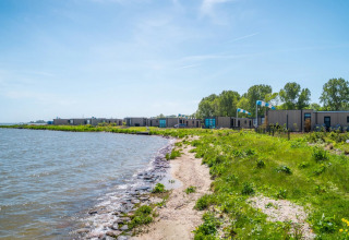 Vakantiepark aan het meer met moderne glampingverblijven, groene natuur en een stralende blauwe lucht.