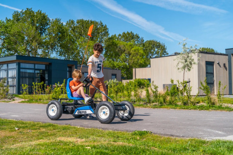 Dos niños juegan con un coche de pedales frente a modernas cabañas glamping en un parque vacacional soleado.