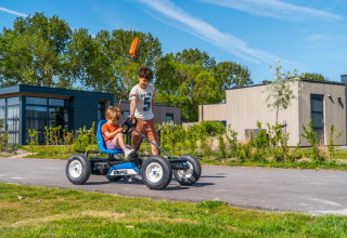 To børn leger med en pedal-gokart foran moderne glamping-hytter i en feriepark på en solrig dag.