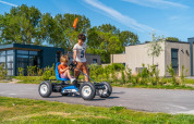 Two children play with a pedal go-kart in front of modern glamping cabins at a holiday park on a sunny day.