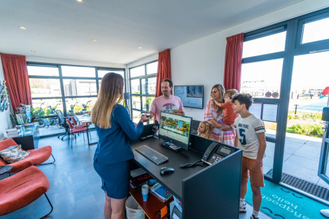 Famille s’enregistre à la réception d’un parc de vacances en glamping, accueillie par la réceptionniste.