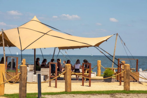Glampingområde med teltskygge, folk sidder ved borde, udsigt til strand og hav under blå himmel.