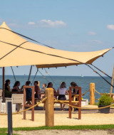 Glampingområde med teltskygge, folk sidder ved borde, udsigt til strand og hav under blå himmel.