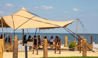 Area glamping con tenda da sole, persone sedute davanti al mare, atmosfera rilassata sulla spiaggia.