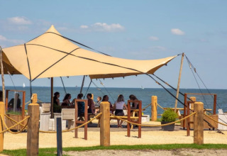 Glampingområde med teltskygge, folk sidder ved borde, udsigt til strand og hav under blå himmel.