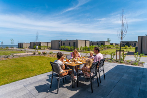 Familie isst im Freien auf einer Terrasse in einem Ferienpark mit modernen Glamping-Unterkünften am Meer.