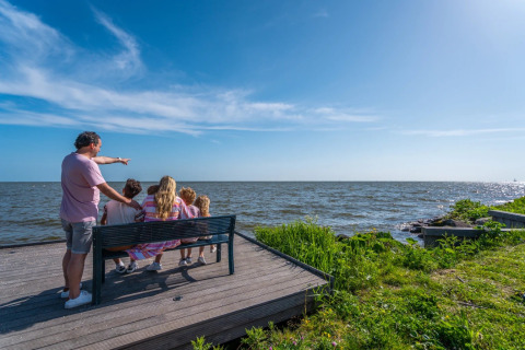 Familie sitzt auf einer Bank am Wasser in einem Ferienpark mit Glamping bei sonnigem Wetter.