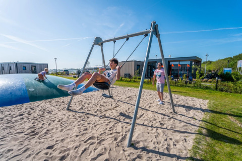 Niños juegan en un columpio y almohadilla de salto en un parque vacacional con alojamientos glamping.
