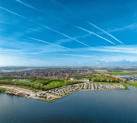 Vista aérea de un parque vacacional junto al lago con alojamientos glamping y cielo azul con estelas blancas.