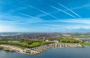 Luftfoto af feriepark ved sø med glampinghytter, grønne områder og blå himmel med kondensstriber.