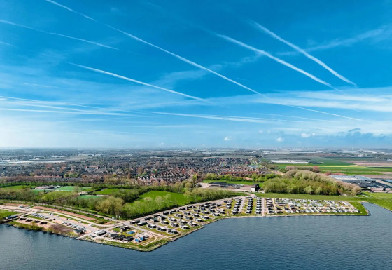 Luchtfoto van een vakantiepark met glamping aan het water, groene zones en een blauwe lucht met strepen.