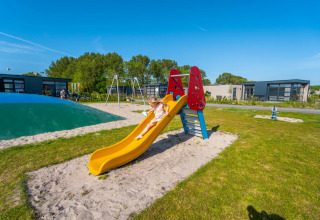Kindje glijdt van een glijbaan op de speeltuin van een glamping vakantiepark met moderne huisjes.