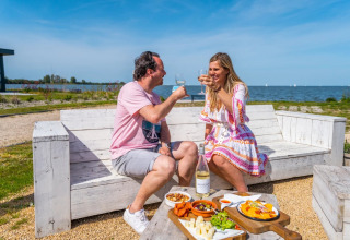 Due persone brindano con vino gustando cibo all’aperto vicino al lago, seduti su panche bianche in un glamping.