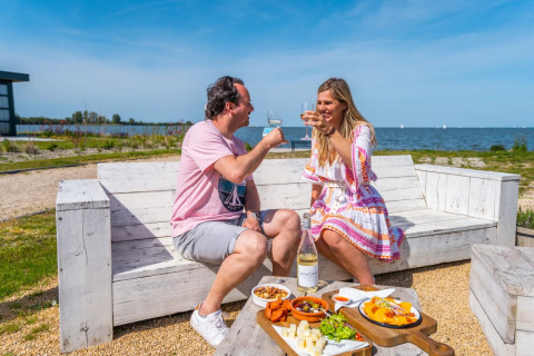 Due persone brindano con vino gustando cibo all’aperto vicino al lago, seduti su panche bianche in un glamping.