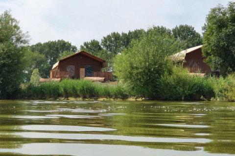 Two glamping cabins by a lakeside or riverbank, surrounded by lush greenery and trees at a holiday park.
