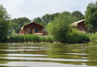 Zwei Glamping-Hütten am Ufer eines Sees oder Flusses, umgeben von üppigem Grün und Bäumen.