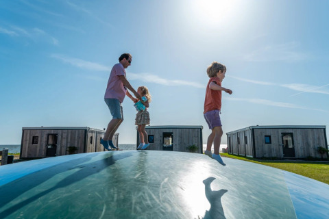 Familia jugando en un gran cojín inflable frente a cabañas glamping en un parque de vacaciones soleado.