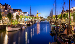 Evening view of a canal lined with sailboats, trees, and charming historic buildings in a Dutch city.