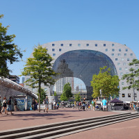 Photo of a busy city square with the modern Markthal building in the background on a sunny day.