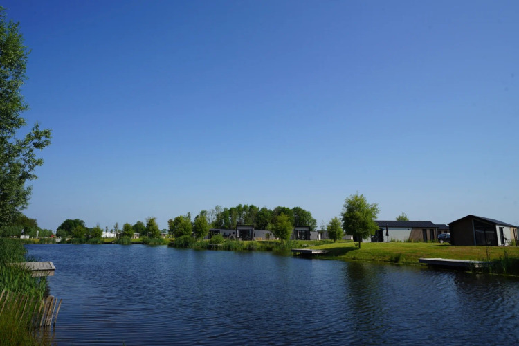Vakantiepark met glamping-accommodaties aan het water, omgeven door bomen, gras en blauwe lucht.