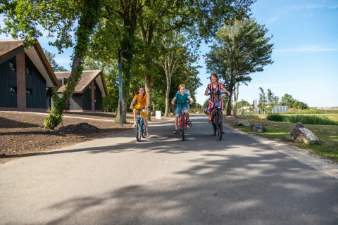 Tre personer cykler fornøjeligt på en solrig dag ved et feriepark med glamping i grønt landskab.