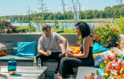 To mennesker nyder en drink på en farverig terrasse ved søen i et feriepark med glamping.