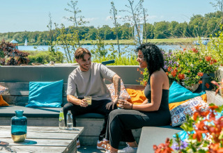 Two people enjoy drinks on a colorful lakefront terrace at a holiday park offering glamping stays.