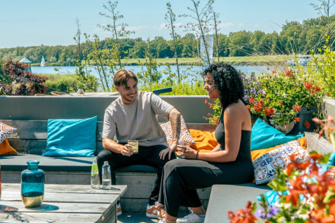 Dos personas disfrutan de bebidas en una terraza junto al lago en un parque vacacional con glamping.