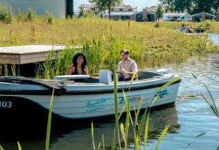 Twee mensen varen in een boot bij een vakantiepark met glampingaccommodaties op de achtergrond.