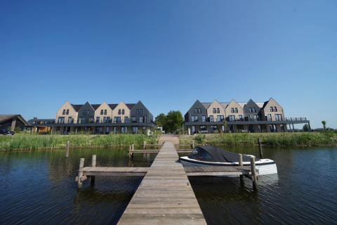 Holiday park with glamping lodges by the water, wooden pier, and boat under a clear blue sky.