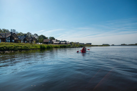 Parque vacacional con cabañas glamping junto al lago, pareja pasea en kayak rojo bajo cielo azul.