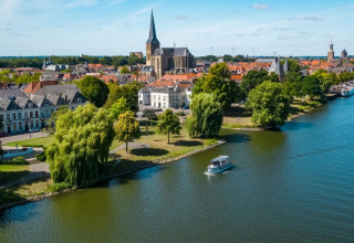 Vue d’une ville au bord d’une rivière avec parcs, bateau et hébergements glamping dans un parc de vacances.