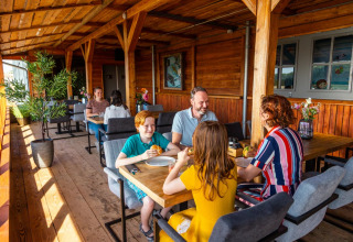 Familie nyder et måltid sammen på en overdækket træterrasse i en feriepark med glamping faciliteter.