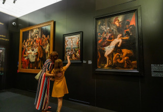 A woman and child admire classic paintings in a museum gallery with large framed artworks on display.
