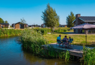 Gezin ontspant op een steiger bij een vijver, omringd door moderne glampinghuisjes in een vakantiepark.