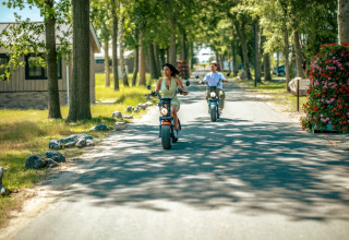 Two people ride electric scooters down a tree-lined road in a holiday park offering glamping accommodations.