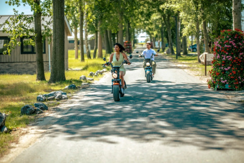 Two people ride electric scooters down a tree-lined road in a holiday park offering glamping accommodations.