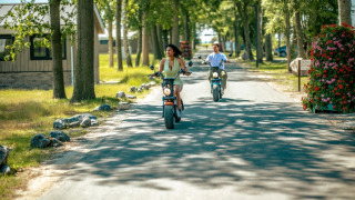 Dos personas montan scooters eléctricos por un camino arbolado en un parque vacacional con tiendas glamping.