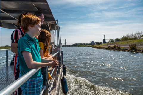 Familie geniet van een boottocht bij een glamping vakantiepark, met uitzicht op water en windmolen.