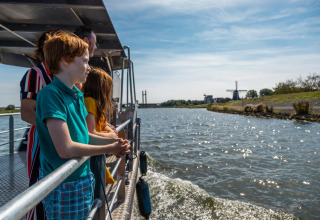 Gezin op boottocht vanuit vakantiepark met glamping, uitkijkend op het water en een windmolen in de verte.