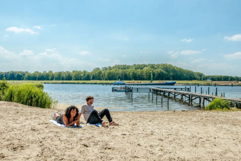 Two people relaxing on a sandy beach by a lake with a wooden pier and forested park in the background.