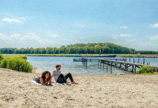 Dos personas descansan en la playa de arena junto a un lago con muelle y bosque en el fondo.