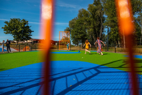 Enfants et adultes jouent sur une aire de jeux colorée avec gazon artificiel dans un parc de vacances, vue à travers une clôture.
