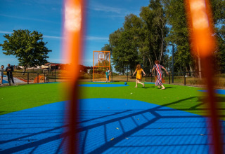 Kinderen en volwassenen spelen op een kleurrijk speelveld met kunstgras in een vakantiepark, gezien door een hek.