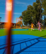Enfants et adultes jouent sur une aire de jeux colorée avec gazon artificiel dans un parc de vacances, vue à travers une clôture.